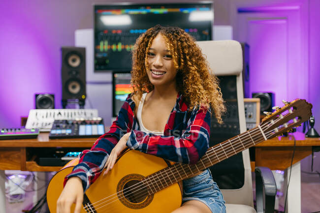 Молодая афроамериканка-гитаристка с классической гитарой сидит в студии и смотрит в камеру — стоковое фото