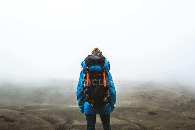 Back view of calm tranquil woman well equipped in bright blue jacket with backpack standing on top and looking at grey valley and mountains in mist — Stock Photo