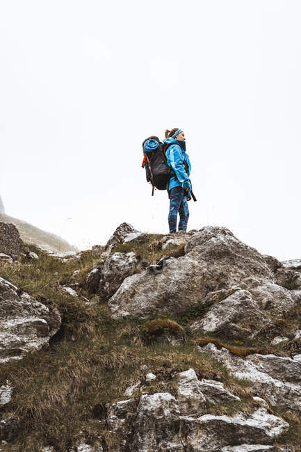 Вид сбоку спокойной женщины в ярко-синей куртке с рюкзаком, стоящей на скальном холме и отводящей взгляд — стоковое фото