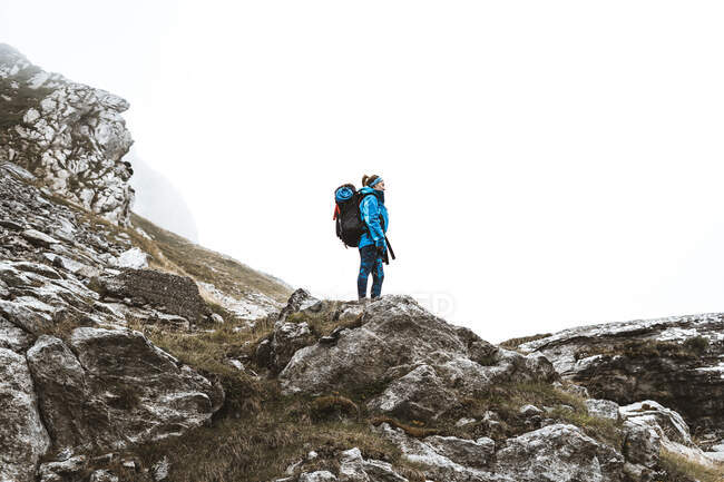 Вид сбоку спокойной женщины в ярко-синей куртке с рюкзаком, стоящей на скальном холме и отводящей взгляд — стоковое фото