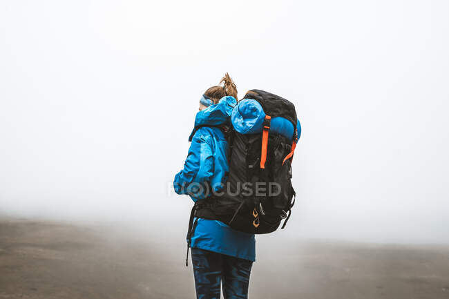Вид збоку спокійна спокійна жінка добре обладнана в яскраво-блакитній куртці з рюкзаком, що стоїть зверху і дивиться на сіру долину і гори в тумані — стокове фото