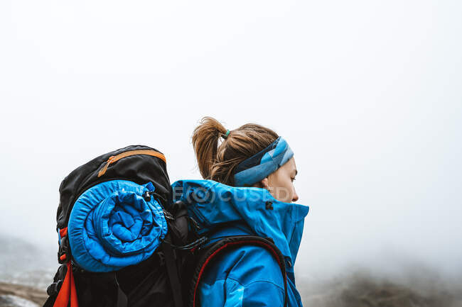Vue latérale de la femme tranquille en veste bleu vif avec sac à dos debout sur une colline rocheuse et regardant loin — Photo de stock
