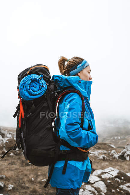 Vista lateral de mujer tranquila en chaqueta azul brillante con mochila de pie en la colina rocosa y mirando hacia otro lado - foto de stock