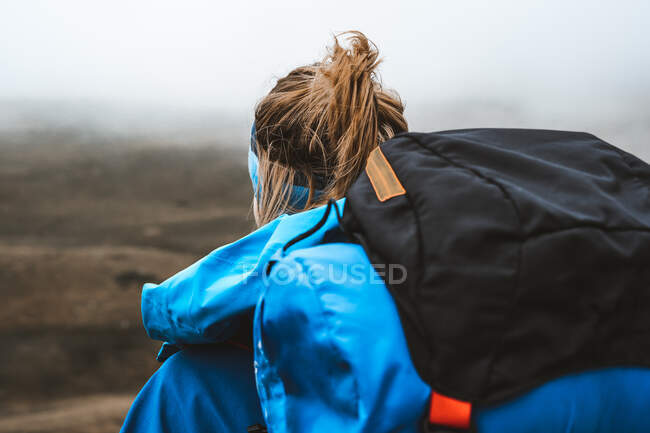 Вид ззаду спокійна спокійна жінка, добре обладнана в яскраво-блакитній куртці з рюкзаком, що стоїть зверху і дивиться на сіру долину і гори в тумані — стокове фото