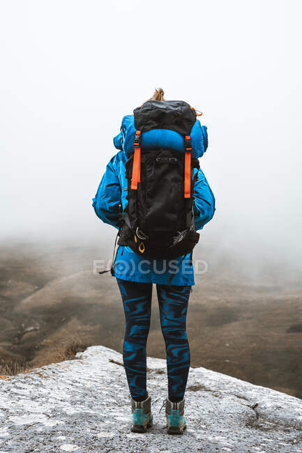 Вид на спокойную женщину в ярко-голубой куртке с рюкзаком, стоящим на скалистом холме и смотрящим в сторону — стоковое фото