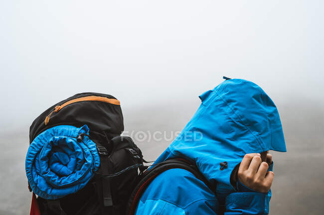 Вид сбоку на неузнаваемую спокойную женщину в ярко-синей куртке, идущую с рюкзаком, смотрящую вниз в сухую долину в туманной дымке — стоковое фото