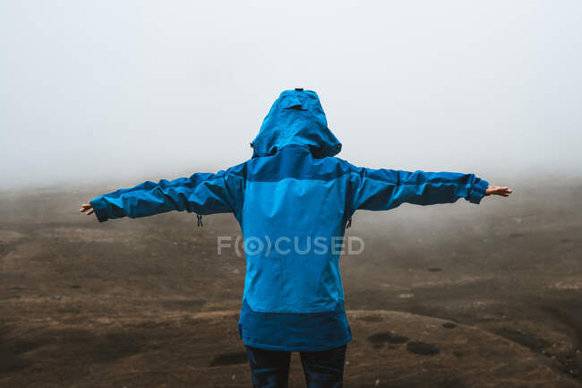 Visão traseira da mulher tranquila irreconhecível em casaco azul brilhante em pé na colina rochosa com os braços abertos — Fotografia de Stock