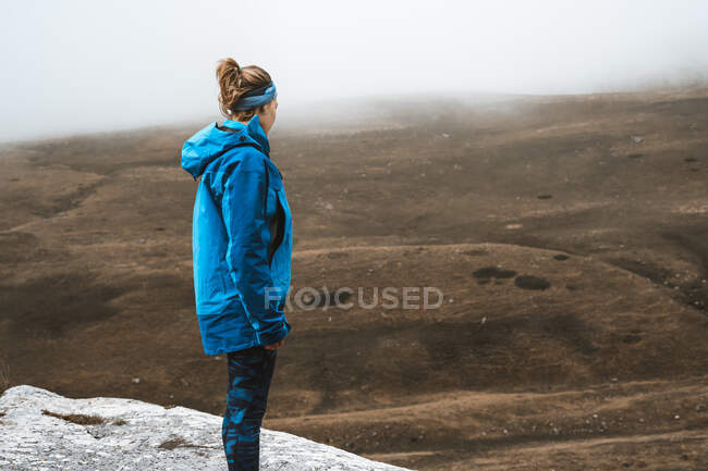 Side view of unrecognizable tranquil woman in bright blue jacket standing on rocky hill — Stock Photo