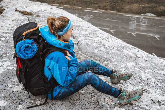 De cima mulher tranquila em casaco azul brilhante com mochila sentada na colina rochosa e olhando para longe — Fotografia de Stock