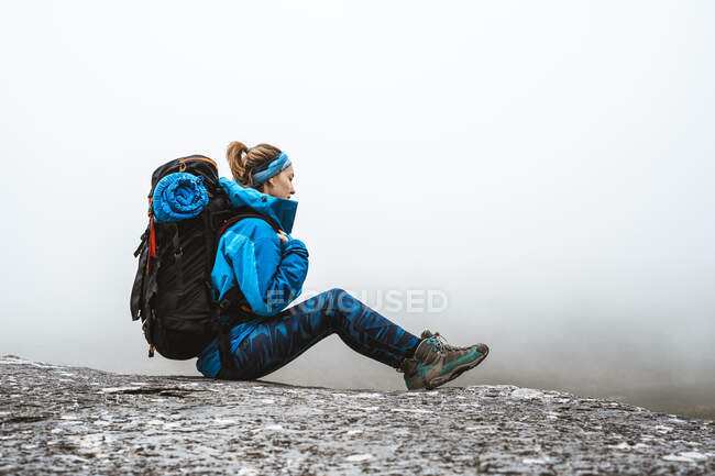 Вид сбоку спокойной женщины в ярко-синей куртке с рюкзаком, сидящей на скальном холме и отводящей взгляд — стоковое фото