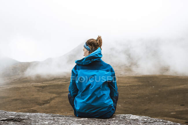 Vista posterior de la mujer tranquila en chaqueta azul brillante con sentado en la colina rocosa y mirando hacia otro lado - foto de stock