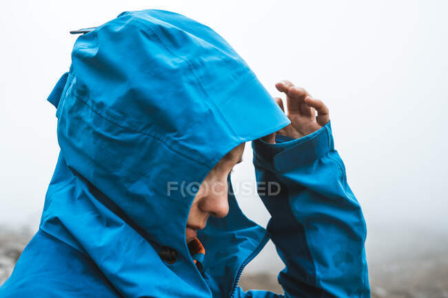 Vista lateral de mujer tranquila irreconocible en chaqueta azul brillante caminando con mochila mirando hacia abajo en valle seco en neblina brumosa - foto de stock