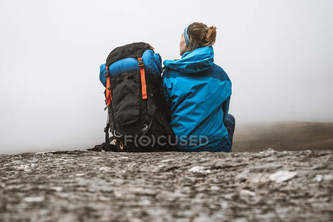 Vista trasera de una mujer tranquila en chaqueta azul brillante con mochila sentada en una colina rocosa y mirando hacia otro lado - foto de stock
