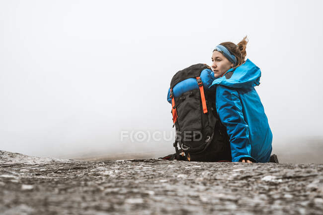 Vista trasera de una mujer tranquila en chaqueta azul brillante con mochila sentada en una colina rocosa y mirando hacia otro lado - foto de stock