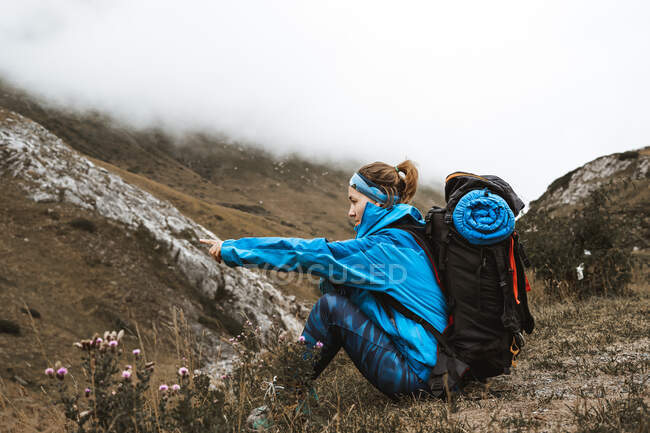 Seitenansicht einer nicht wiederzuerkennenden, ruhigen Frau in hellblauer Jacke, die sich mit ihrem Rucksack ausruht und den Blick im nebelverhangenen Tal genießt — Stockfoto
