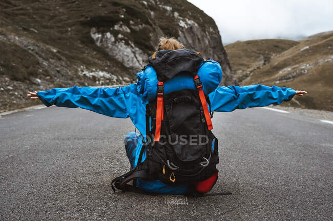 Вид ззаду спокійна жінка в яскраво-синій куртці з рюкзаком сидить на дорозі з відкритими руками в гірському пейзажі — стокове фото