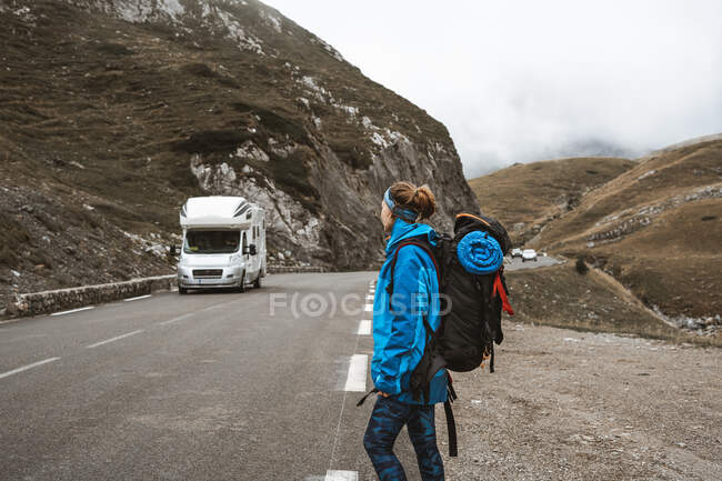 Vista laterale della donna che viaggia con grande zaino e in giacca blu su strada in attesa veicolo bianco — Foto stock