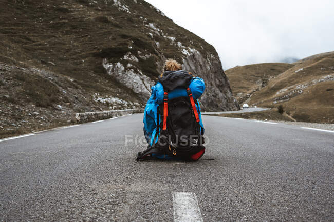 Visão traseira da mulher tranquila em casaco azul brilhante com mochila sentada na estrada e olhando para longe — Fotografia de Stock