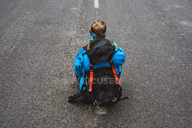 De cima visão traseira da mulher tranquila em casaco azul brilhante com mochila sentada na estrada e olhando para longe — Fotografia de Stock