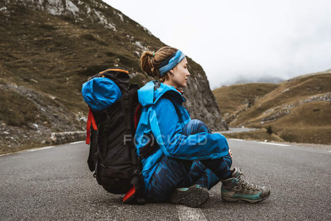 Vista lateral de mujer tranquila en chaqueta azul brillante con mochila sentada en la carretera y mirando hacia otro lado - foto de stock