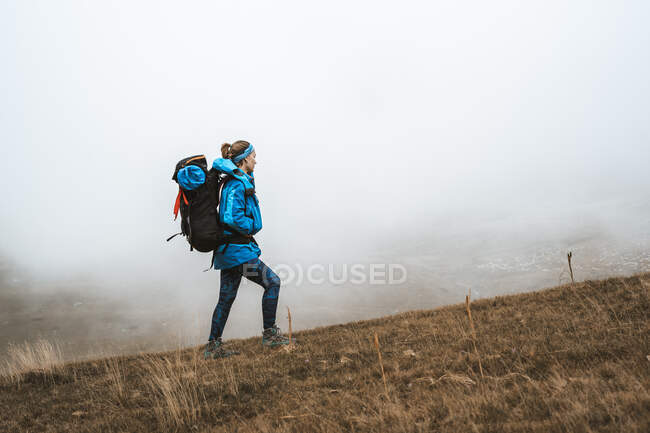 Vista lateral da mulher tranquila em casaco azul brilhante e mochila em pé no vale seco em neblina nebulosa olhando para longe — Fotografia de Stock