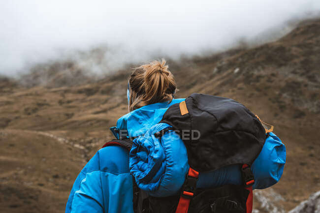 Rückansicht einer ruhigen Frau in hellblauer Jacke mit Rucksack, die auf einem felsigen Hügel steht und wegschaut — Stockfoto