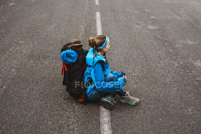 De cima vista lateral da mulher tranquila em casaco azul brilhante com mochila sentada na estrada e olhando para longe — Fotografia de Stock