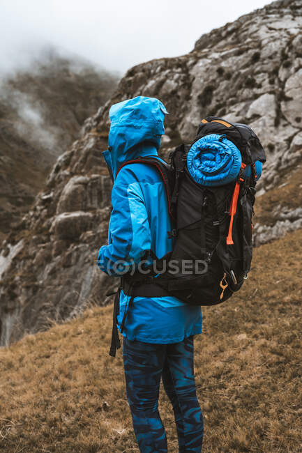 Vista lateral da mulher anônima tranquila em casaco azul brilhante com mochila em pé na colina rochosa e olhando para longe — Fotografia de Stock
