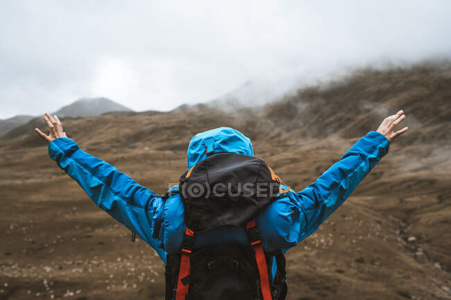 Vista trasera de una mujer tranquila irreconocible con chaqueta azul brillante de pie en una colina rocosa con los brazos abiertos - foto de stock