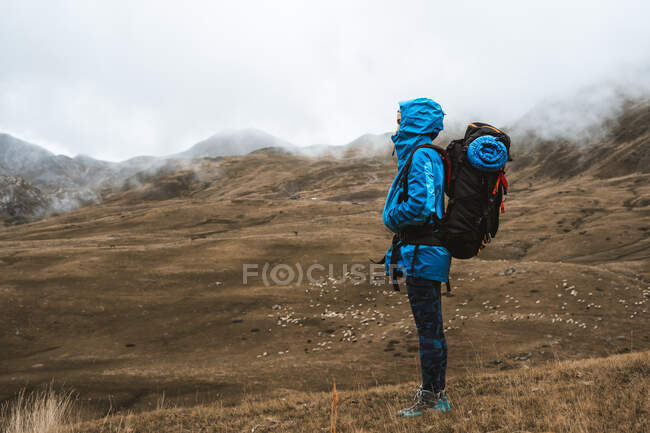 Vista lateral de mujer tranquila en chaqueta azul brillante con sudadera con capucha y mochila de pie en valle seco en neblina brumosa - foto de stock