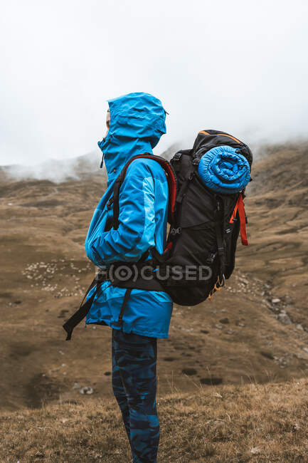 Vista lateral de la tranquila mujer anónima en chaqueta azul brillante con mochila de pie en la colina rocosa y mirando hacia otro lado - foto de stock