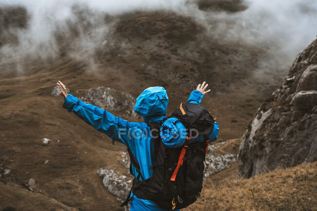 Back view of unrecognizable tranquil woman in bright blue jacket standing on rocky hill with open arms — Stock Photo