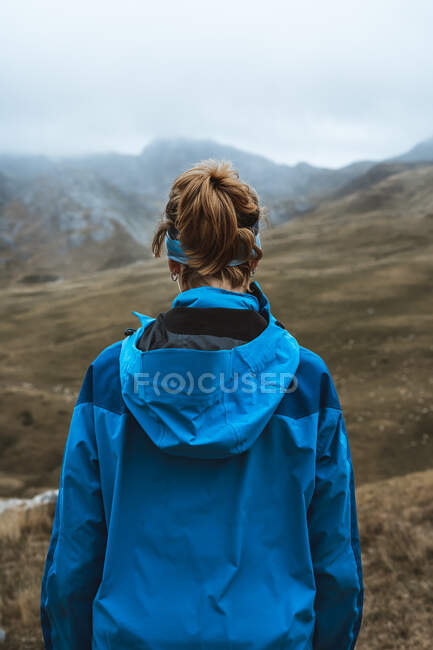 Вид на спокойную женщину в ярко-голубой куртке с рюкзаком, стоящим на скалистом холме и смотрящим в сторону — стоковое фото