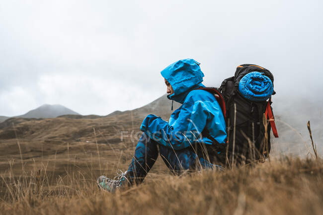 Vista lateral de mujer tranquila irreconocible en chaqueta azul brillante descansando sentada con mochila disfrutando de vistas mirando hacia otro lado en valle seco en neblina - foto de stock