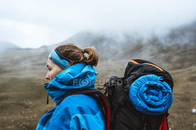 Side view of tranquil anonymous woman in bright blue jacket with backpack standing on rocky hill and looking away — Stock Photo