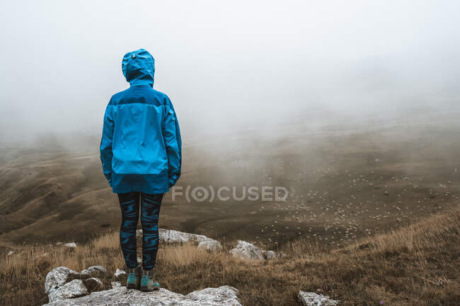Visão traseira da mulher tranquila irreconhecível em casaco azul brilhante em pé na colina rochosa — Fotografia de Stock