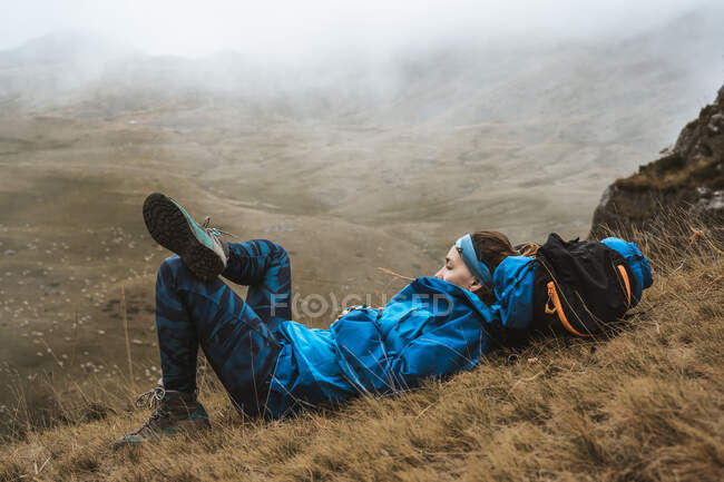 Side view of tranquil woman in bright blue jacket having rest leaning on backpack and lying with crossed leg in dry valley in foggy haze — Stock Photo