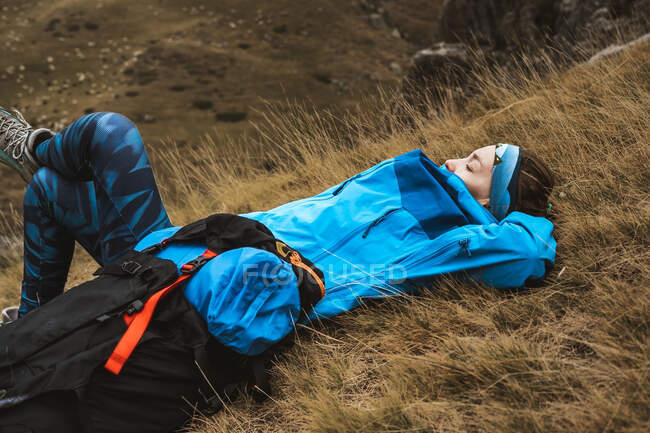 Vista lateral de mujer tranquila en chaqueta azul brillante que descansa apoyada en la mochila y acostada con la pierna cruzada en valle seco en neblina - foto de stock