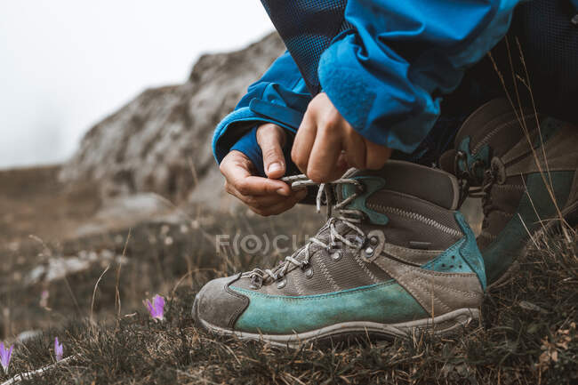 Crop person in blue jacket crouching and tying shoelaces on boot on way in mountains — Stock Photo
