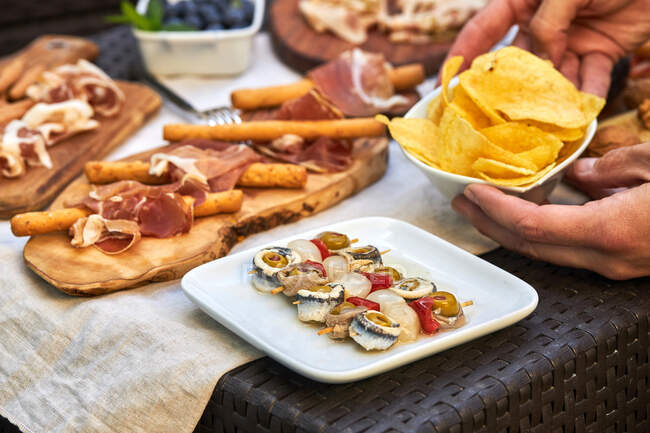 Unknown hands picking up a bowl of potato chips from a table full of various snacks such as skewered pickled, serrano ham, nuts, cheese, etc.unknown hand picking up serrano ham with bread on a table filled with various snacks such as spicy banderitas — Stock Photo