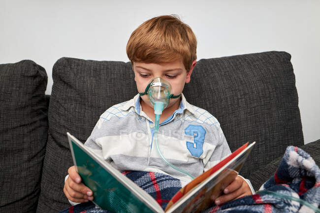 Boy in oxygen mask using nebulizer during inhalation while sitting on sofa and reading book — Stock Photo