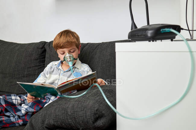 Junge in Sauerstoffmaske mit Vernebler beim Inhalieren, während er auf Sofa sitzt und Buch liest — Stockfoto