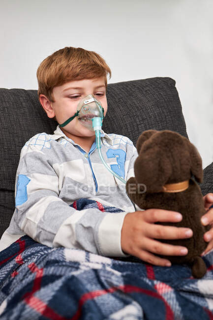 Enfant joyeux respirant masque à oxygène pendant l'inhalation et jouant avec un jouet doux sur le canapé à la maison — Photo de stock