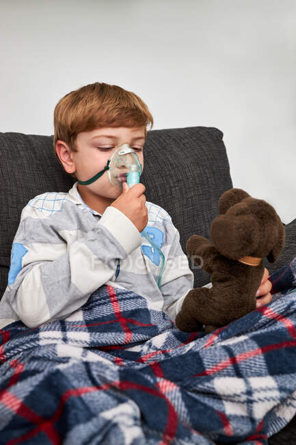 Enfant joyeux respirant masque à oxygène pendant l'inhalation et jouant avec un jouet doux sur le canapé à la maison — Photo de stock