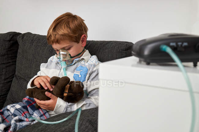 Alegre niño respirando la máscara de oxígeno durante la inhalación y jugando con el juguete suave en el sofá en casa - foto de stock