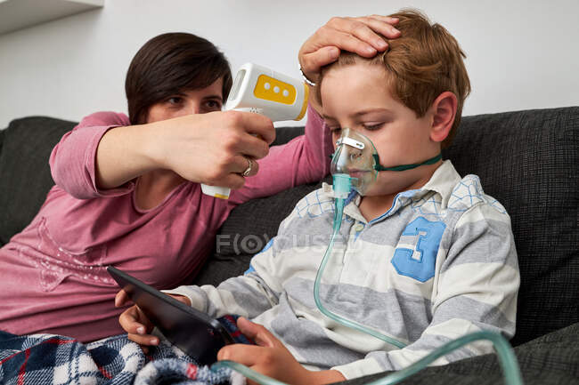 Madre amorevole con termometro a infrarossi che misura la temperatura del ragazzo respirando maschera di ossigeno durante la procedura di inalazione e guardando cartone animato su tablet a casa — Foto stock