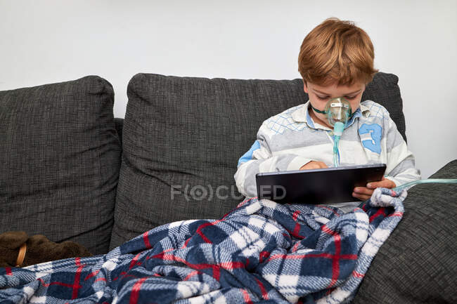 Garçon malade en masque à oxygène assis sur le canapé à la maison et jouer à un jeu vidéo sur tablette — Photo de stock