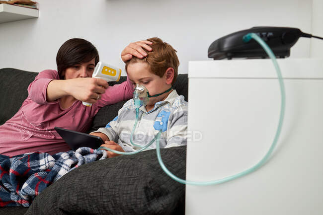 Mãe amorosa com termômetro infravermelho medindo a temperatura do menino respirando máscara de oxigênio durante o procedimento de inalação e assistindo desenhos animados em comprimido em casa — Fotografia de Stock