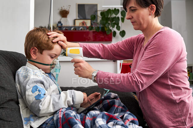 Mère aimante avec thermomètre infrarouge mesurant la température du garçon respirant masque à oxygène pendant la procédure d'inhalation et regardant bande dessinée sur tablette à la maison — Photo de stock