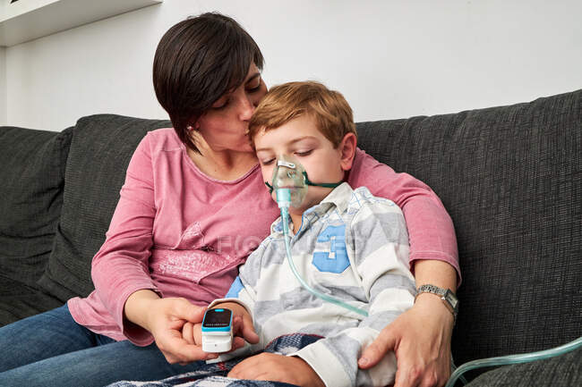 Donna premurosa che utilizza pulsossimetro al dito del ragazzo malato in maschera di ossigeno utilizzando nebulizzatore durante l'inalazione a casa — Foto stock